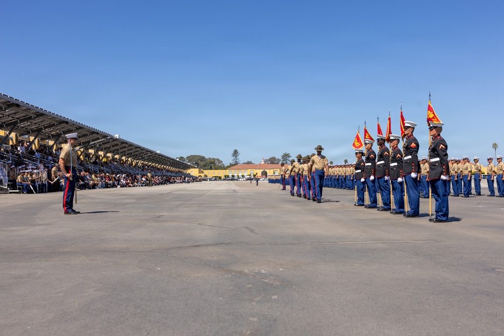 A formal U.S. Marine Corps graduation or pass-in-review ceremony is taking place on a large parade deck. Marines in dress blue uniforms are standing in formation holding guidon flags, while others in service khaki uniforms observe or command. A large grandstand filled with spectators — likely family members — lines the left side of the parade field.