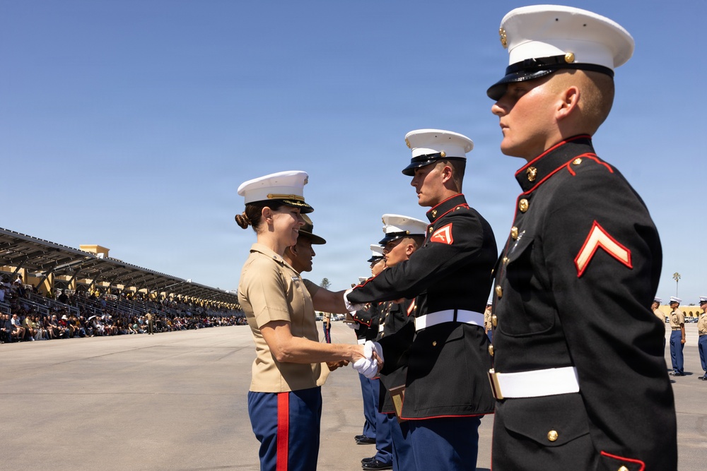 A U.S. Marine Corps graduation or awards ceremony is taking place on a parade deck, likely at MCRD San Diego based on the visible bleachers and facility layout. A female officer in service khaki uniform is shaking hands with and presenting something to Marines in dress blue uniforms. Spectators fill the bleachers in the background, indicating a formal public ceremony.