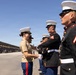 A U.S. Marine Corps graduation or awards ceremony is taking place on a parade deck, likely at MCRD San Diego based on the visible bleachers and facility layout. A female officer in service khaki uniform is shaking hands with and presenting something to Marines in dress blue uniforms. Spectators fill the bleachers in the background, indicating a formal public ceremony.
