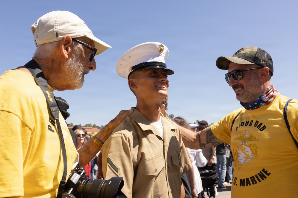 A newly graduated U.S. Marine in dress uniform is being congratulated by two older men at what appears to be a military graduation ceremony. The man on the right is wearing a yellow 'Proud Dad U.S. Marine' shirt and has his hand on the Marine's shoulder. The setting appears to be outdoors at a military base with a crowd visible in the background.