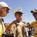 A newly graduated U.S. Marine in dress uniform is being congratulated by two older men at what appears to be a military graduation ceremony. The man on the right is wearing a yellow 'Proud Dad U.S. Marine' shirt and has his hand on the Marine's shoulder. The setting appears to be outdoors at a military base with a crowd visible in the background.