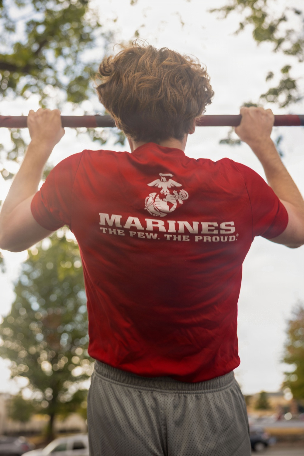 A person is performing pull-ups on an outdoor bar, viewed from behind. They are wearing a red U.S. Marines t-shirt displaying the Eagle, Globe, and Anchor emblem along with the slogan 'The Few. The Proud.' The background shows trees and a partially overcast sky in an outdoor setting.