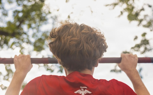 A person is performing pull-ups on an outdoor bar, viewed from behind. They are wearing a red U.S. Marines t-shirt displaying the Eagle, Globe, and Anchor emblem along with the slogan 'The Few. The Proud.' The background shows trees and a partially overcast sky in an outdoor setting.