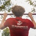 A person is performing pull-ups on an outdoor bar, viewed from behind. They are wearing a red U.S. Marines t-shirt displaying the Eagle, Globe, and Anchor emblem along with the slogan 'The Few. The Proud.' The background shows trees and a partially overcast sky in an outdoor setting.