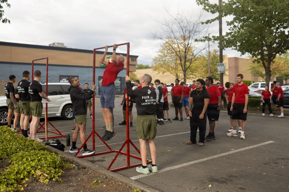 A group of individuals in athletic wear are gathered in a parking lot for what appears to be a military recruiting or fitness event. A person in a red shirt is performing pull-ups on a portable red pull-up bar frame while others observe. The crowd consists of both instructors in black shirts and participants in red shirts, suggesting a structured fitness assessment or demonstration.