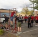 A group of individuals in athletic wear are gathered in a parking lot for what appears to be a military recruiting or fitness event. A person in a red shirt is performing pull-ups on a portable red pull-up bar frame while others observe. The crowd consists of both instructors in black shirts and participants in red shirts, suggesting a structured fitness assessment or demonstration.