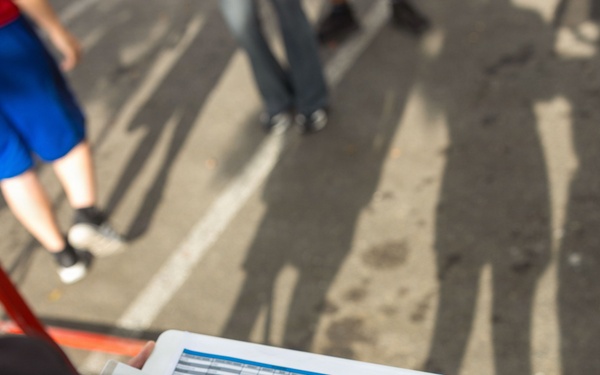 A tattooed individual is writing on a clipboard with a structured form/spreadsheet attached to it outdoors. The clipboard features a logo that partially reads 'MA...ES' in white text on a navy and red background, possibly a sports team or organization. The scene appears to take place on a street or parking area with other people visible in the background.