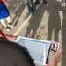 A tattooed individual is writing on a clipboard with a structured form/spreadsheet attached to it outdoors. The clipboard features a logo that partially reads 'MA...ES' in white text on a navy and red background, possibly a sports team or organization. The scene appears to take place on a street or parking area with other people visible in the background.