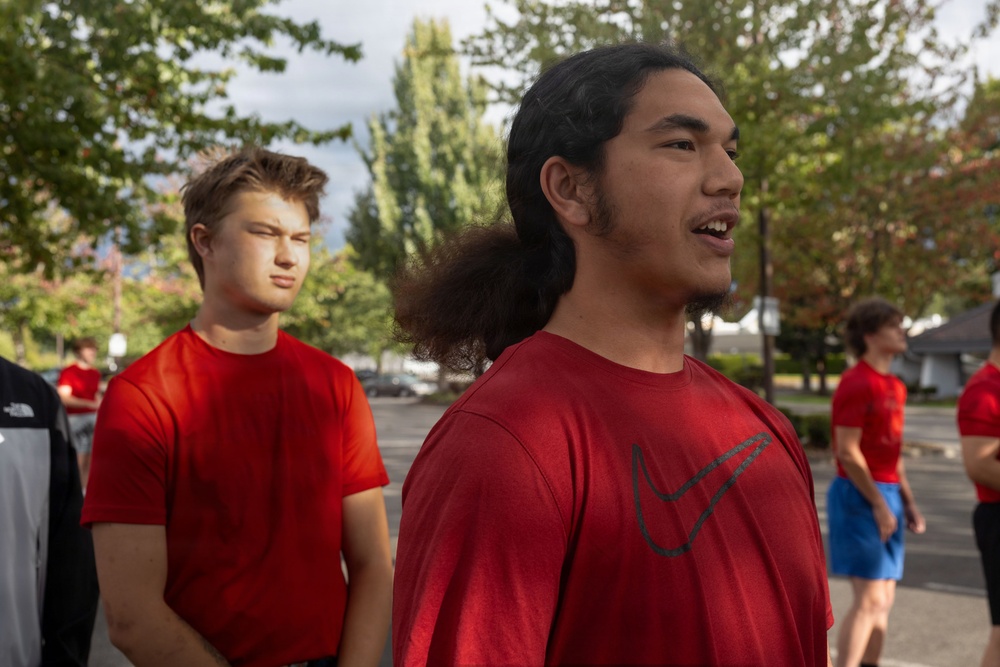 A group of young men wearing matching red t-shirts are gathered outdoors in what appears to be a parking lot or open area surrounded by trees. The foreground features two young men prominently, one wearing a red Nike t-shirt with long dark hair tied back, appearing to be speaking. The setting and matching attire suggest a team or group activity, possibly a sports practice or organized event.