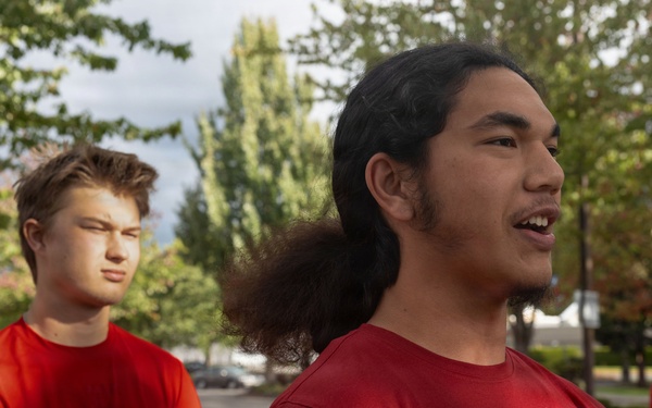A group of young men wearing matching red t-shirts are gathered outdoors in what appears to be a parking lot or open area surrounded by trees. The foreground features two young men prominently, one wearing a red Nike t-shirt with long dark hair tied back, appearing to be speaking. The setting and matching attire suggest a team or group activity, possibly a sports practice or organized event.
