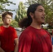 A group of young men wearing matching red t-shirts are gathered outdoors in what appears to be a parking lot or open area surrounded by trees. The foreground features two young men prominently, one wearing a red Nike t-shirt with long dark hair tied back, appearing to be speaking. The setting and matching attire suggest a team or group activity, possibly a sports practice or organized event.