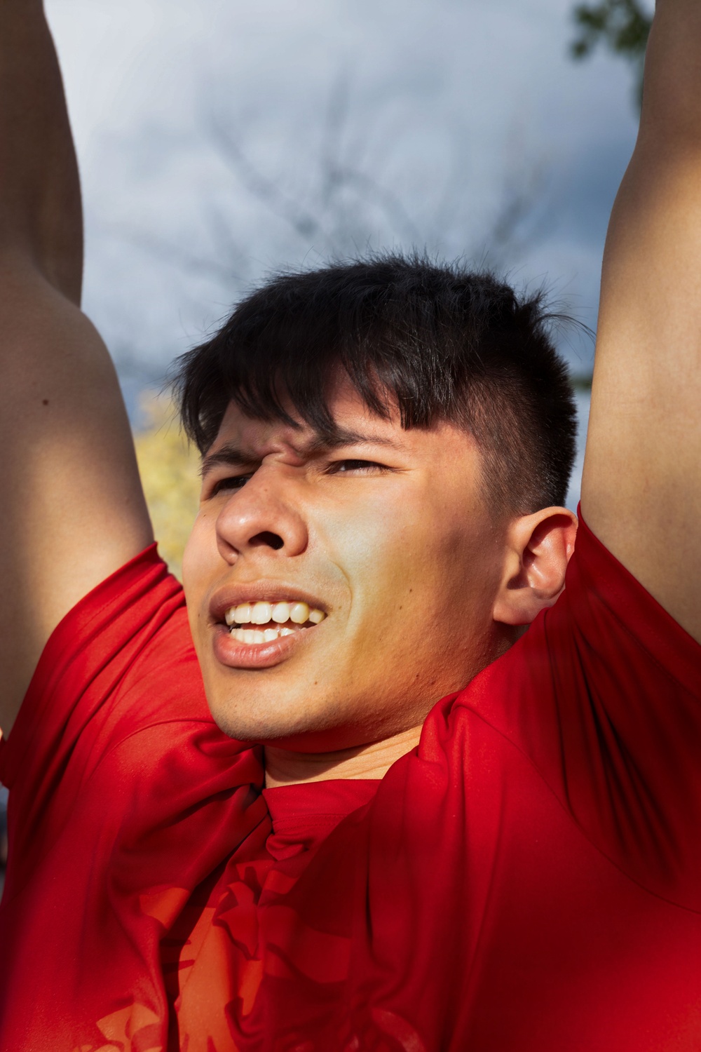 A young man wearing a red athletic shirt is photographed from a low angle with his arms raised above his head. He appears to be outdoors, squinting and grimacing slightly, possibly due to sunlight or physical exertion. The background shows a partly cloudy sky and bare tree branches.