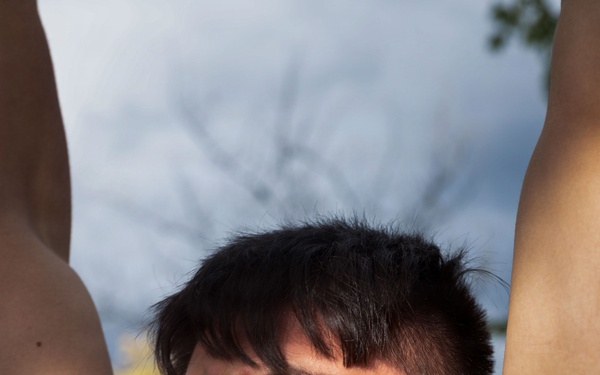 A young man wearing a red athletic shirt is photographed from a low angle with his arms raised above his head. He appears to be outdoors, squinting and grimacing slightly, possibly due to sunlight or physical exertion. The background shows a partly cloudy sky and bare tree branches.