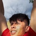 A young man wearing a red athletic shirt is photographed from a low angle with his arms raised above his head. He appears to be outdoors, squinting and grimacing slightly, possibly due to sunlight or physical exertion. The background shows a partly cloudy sky and bare tree branches.