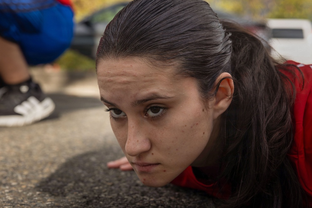 A young woman with dark hair pulled back is positioned very close to the ground, appearing to be performing a push-up or low crawl exercise on pavement. Her expression is focused and determined. A second person in blue clothing and black sneakers is visible in the blurred background.