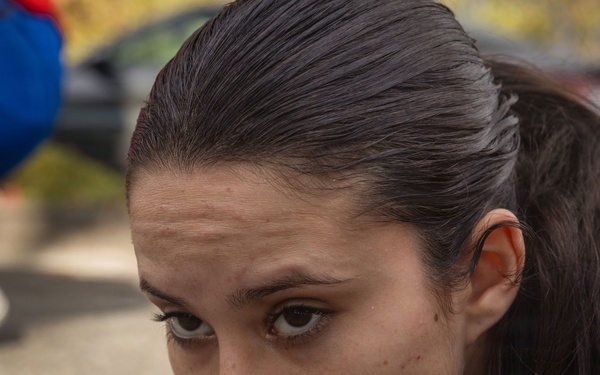 A young woman with dark hair pulled back is positioned very close to the ground, appearing to be performing a push-up or low crawl exercise on pavement. Her expression is focused and determined. A second person in blue clothing and black sneakers is visible in the blurred background.