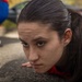 A young woman with dark hair pulled back is positioned very close to the ground, appearing to be performing a push-up or low crawl exercise on pavement. Her expression is focused and determined. A second person in blue clothing and black sneakers is visible in the blurred background.