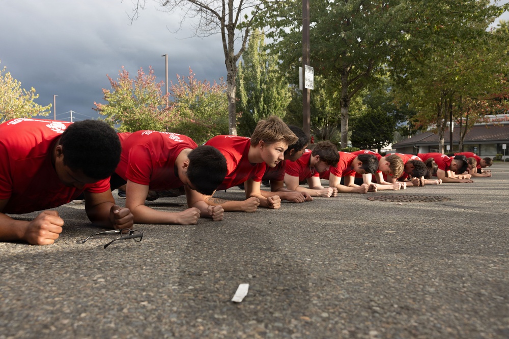 A large group of young males wearing matching red t-shirts are performing a synchronized plank exercise in a parking lot. They are aligned in a single row extending into the background, suggesting a team or group training session. The setting appears to be an outdoor paved area with trees and overcast skies visible in the background.
