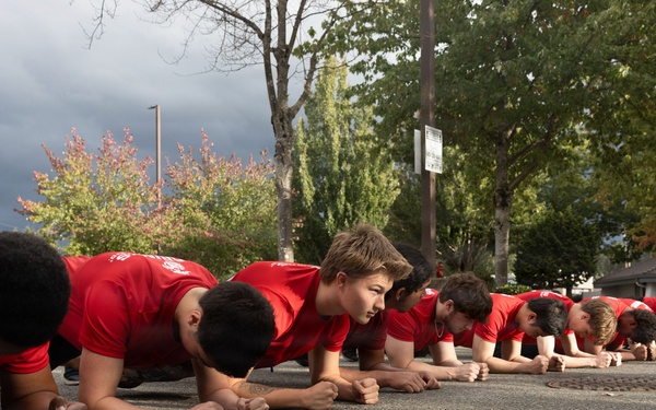 A large group of young males wearing matching red t-shirts are performing a synchronized plank exercise in a parking lot. They are aligned in a single row extending into the background, suggesting a team or group training session. The setting appears to be an outdoor paved area with trees and overcast skies visible in the background.