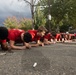 A large group of young males wearing matching red t-shirts are performing a synchronized plank exercise in a parking lot. They are aligned in a single row extending into the background, suggesting a team or group training session. The setting appears to be an outdoor paved area with trees and overcast skies visible in the background.