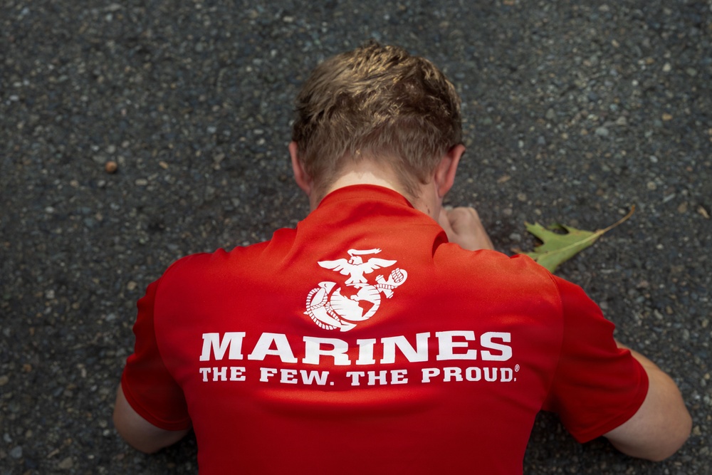 An overhead view of a person wearing a red U.S. Marines branded t-shirt, photographed from above while they appear to be crouching or leaning forward on a paved surface. The shirt displays the Eagle, Globe, and Anchor emblem along with the text 'Marines - The Few. The Proud.' A fallen leaf is visible near the person's shoulder.