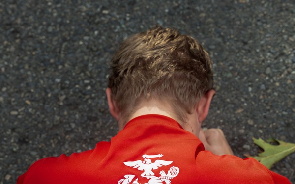 An overhead view of a person wearing a red U.S. Marines branded t-shirt, photographed from above while they appear to be crouching or leaning forward on a paved surface. The shirt displays the Eagle, Globe, and Anchor emblem along with the text 'Marines - The Few. The Proud.' A fallen leaf is visible near the person's shoulder.