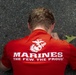An overhead view of a person wearing a red U.S. Marines branded t-shirt, photographed from above while they appear to be crouching or leaning forward on a paved surface. The shirt displays the Eagle, Globe, and Anchor emblem along with the text 'Marines - The Few. The Proud.' A fallen leaf is visible near the person's shoulder.