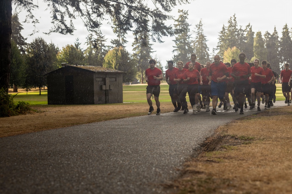 A large group of individuals wearing matching red t-shirts and dark shorts are running together in formation along a paved path, suggesting a military or organized physical training exercise. The setting appears to be a military base or institutional grounds, featuring manicured grass, tall evergreen trees, and a small wooden utility shed. The scene is captured in wet, overcast conditions with visible rain or mist in the air.