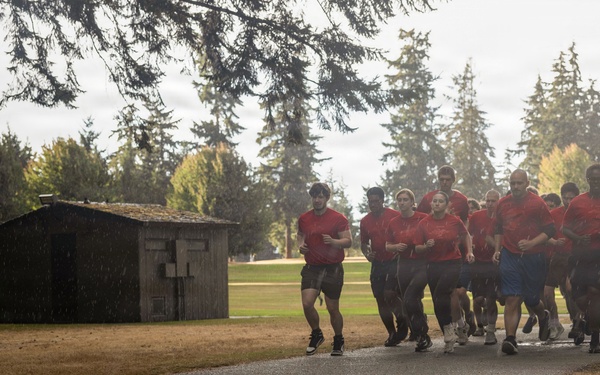A large group of individuals wearing matching red t-shirts and dark shorts are running together in formation along a paved path, suggesting a military or organized physical training exercise. The setting appears to be a military base or institutional grounds, featuring manicured grass, tall evergreen trees, and a small wooden utility shed. The scene is captured in wet, overcast conditions with visible rain or mist in the air.