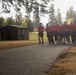 A large group of individuals wearing matching red t-shirts and dark shorts are running together in formation along a paved path, suggesting a military or organized physical training exercise. The setting appears to be a military base or institutional grounds, featuring manicured grass, tall evergreen trees, and a small wooden utility shed. The scene is captured in wet, overcast conditions with visible rain or mist in the air.