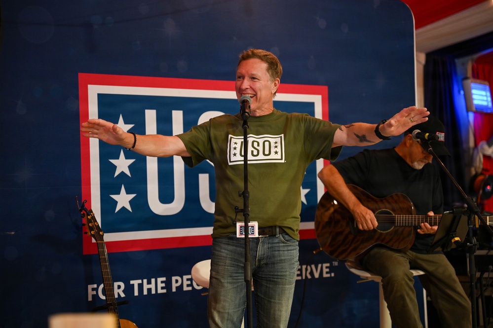 A man wearing a green USO t-shirt performs on stage at a USO event, standing at a microphone with arms outstretched in an engaging gesture. Behind him is a large USO banner reading 'FOR THE PEOPLE WE SERVE.' A second performer sits to his right playing an acoustic guitar.