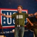 A man wearing a green USO t-shirt performs on stage at a USO event, standing at a microphone with arms outstretched in an engaging gesture. Behind him is a large USO banner reading 'FOR THE PEOPLE WE SERVE.' A second performer sits to his right playing an acoustic guitar.
