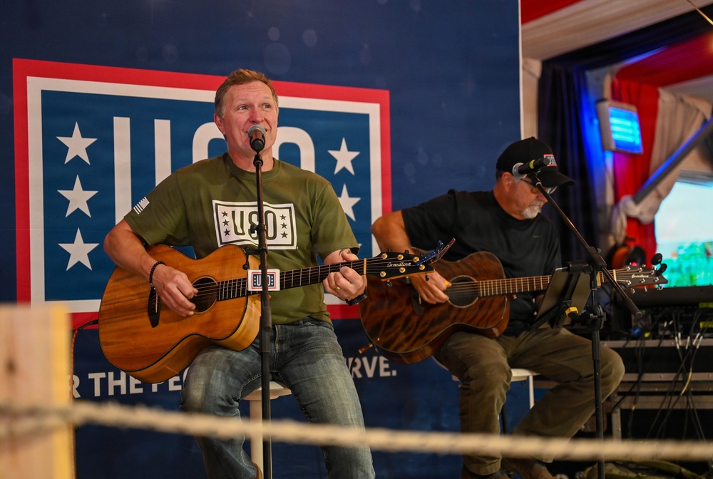 Two musicians are performing at a USO event, with the primary performer singing into a microphone while playing an acoustic guitar and wearing a green USO-branded t-shirt. A second guitarist is seated behind him, also playing an acoustic guitar. The backdrop prominently displays the USO logo with stars and partial text reading 'for the people who serve.'