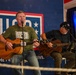 Two musicians are performing at a USO event, with the primary performer singing into a microphone while playing an acoustic guitar and wearing a green USO-branded t-shirt. A second guitarist is seated behind him, also playing an acoustic guitar. The backdrop prominently displays the USO logo with stars and partial text reading 'for the people who serve.'
