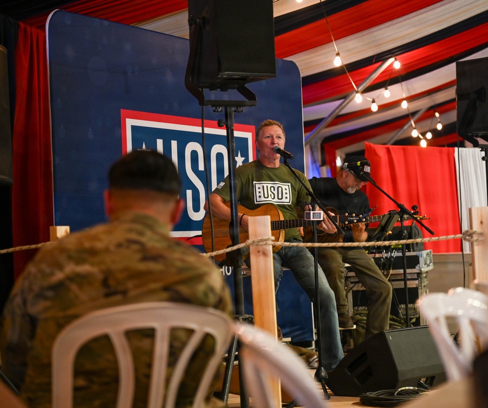 A USO entertainment event is taking place on a small stage where two musicians are performing with acoustic guitars. A military service member in camouflage uniform is seated in the foreground watching the performance. The venue is decorated with red, white, and blue draping and string lights, consistent with a patriotic military support event.