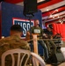 A USO entertainment event is taking place on a small stage where two musicians are performing with acoustic guitars. A military service member in camouflage uniform is seated in the foreground watching the performance. The venue is decorated with red, white, and blue draping and string lights, consistent with a patriotic military support event.