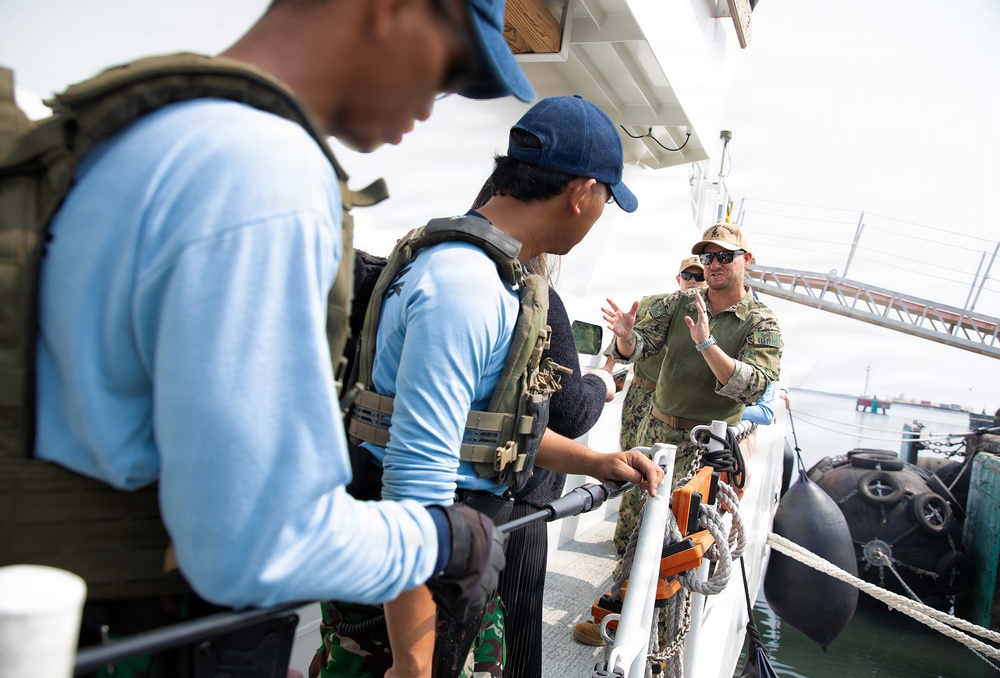 The image shows a group of military or law enforcement personnel on the deck of a vessel, appearing to be engaged in a briefing or training exercise. At least one individual is wearing camouflage military uniform while others wear tactical vests over civilian-style clothing. The setting appears to be a harbor or port environment with fenders and rigging visible along the vessel's side.