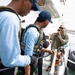 The image shows a group of military or law enforcement personnel on the deck of a vessel, appearing to be engaged in a briefing or training exercise. At least one individual is wearing camouflage military uniform while others wear tactical vests over civilian-style clothing. The setting appears to be a harbor or port environment with fenders and rigging visible along the vessel's side.