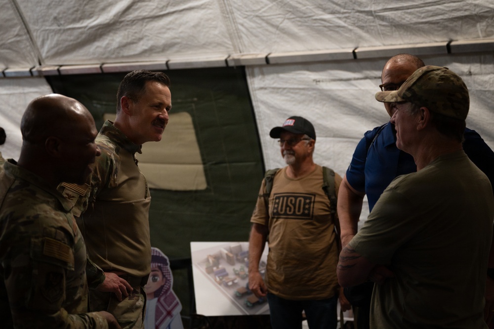 A group of five individuals are gathered inside a military tent structure, engaged in conversation. Two individuals are wearing military camouflage uniforms, while others appear to be civilians, one visibly wearing a USO t-shirt. The setting suggests a USO visit or morale event at a military installation.