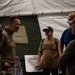 A group of five individuals are gathered inside a military tent structure, engaged in conversation. Two individuals are wearing military camouflage uniforms, while others appear to be civilians, one visibly wearing a USO t-shirt. The setting suggests a USO visit or morale event at a military installation.