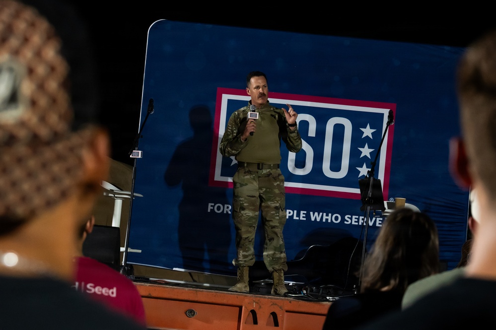 A military service member in camouflage uniform stands on a raised platform speaking into a microphone at a USO event. The speaker is addressing an audience visible in the foreground, with a large USO backdrop behind them reading 'For the People Who Serve.' The setting appears to be an indoor or hangar-type venue with low ambient lighting.