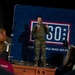 A military service member in camouflage uniform stands on a raised platform speaking into a microphone at a USO event. The speaker is addressing an audience visible in the foreground, with a large USO backdrop behind them reading 'For the People Who Serve.' The setting appears to be an indoor or hangar-type venue with low ambient lighting.