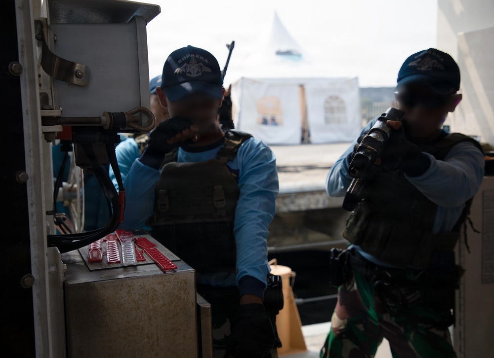 Two armed personnel wearing tactical vests and blue caps are positioned near what appears to be a vessel or dock structure. Both individuals have their faces obscured/blurred, and one is holding a firearm while the other appears to be communicating via radio or similar device. A white tent structure is visible in the background.
