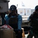 Two armed personnel wearing tactical vests and blue caps are positioned near what appears to be a vessel or dock structure. Both individuals have their faces obscured/blurred, and one is holding a firearm while the other appears to be communicating via radio or similar device. A white tent structure is visible in the background.