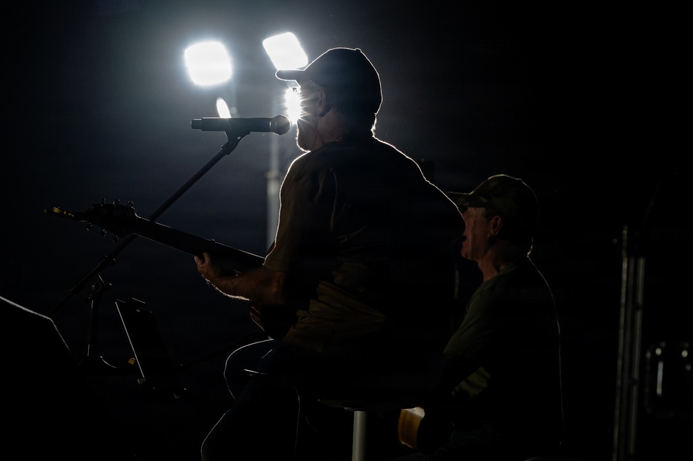 A dramatic backlit photograph of two musicians performing on a dark stage. The primary figure is seated, playing an electric guitar and singing into a microphone, while a second figure wearing a cap is visible in the background. Strong stage lighting from behind creates a silhouette effect, highlighting the performers' outlines against a dark background.