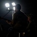 A dramatic backlit photograph of two musicians performing on a dark stage. The primary figure is seated, playing an electric guitar and singing into a microphone, while a second figure wearing a cap is visible in the background. Strong stage lighting from behind creates a silhouette effect, highlighting the performers' outlines against a dark background.