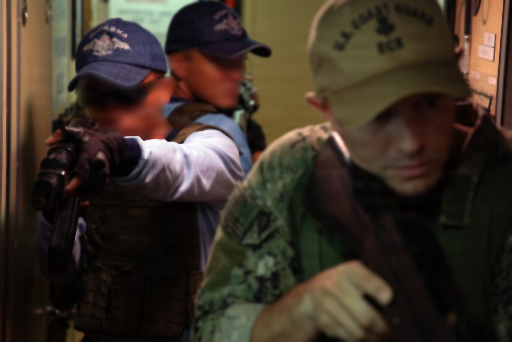 The image shows what appears to be a law enforcement or military boarding/clearance operation in a confined corridor or passageway. Multiple personnel are visible, with at least one individual aiming a firearm and others wearing tactical vests and baseball caps. The foreground figure wears a cap with 'U.S. Coast Guard' text visible.