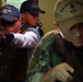 The image shows what appears to be a law enforcement or military boarding/clearance operation in a confined corridor or passageway. Multiple personnel are visible, with at least one individual aiming a firearm and others wearing tactical vests and baseball caps. The foreground figure wears a cap with 'U.S. Coast Guard' text visible.
