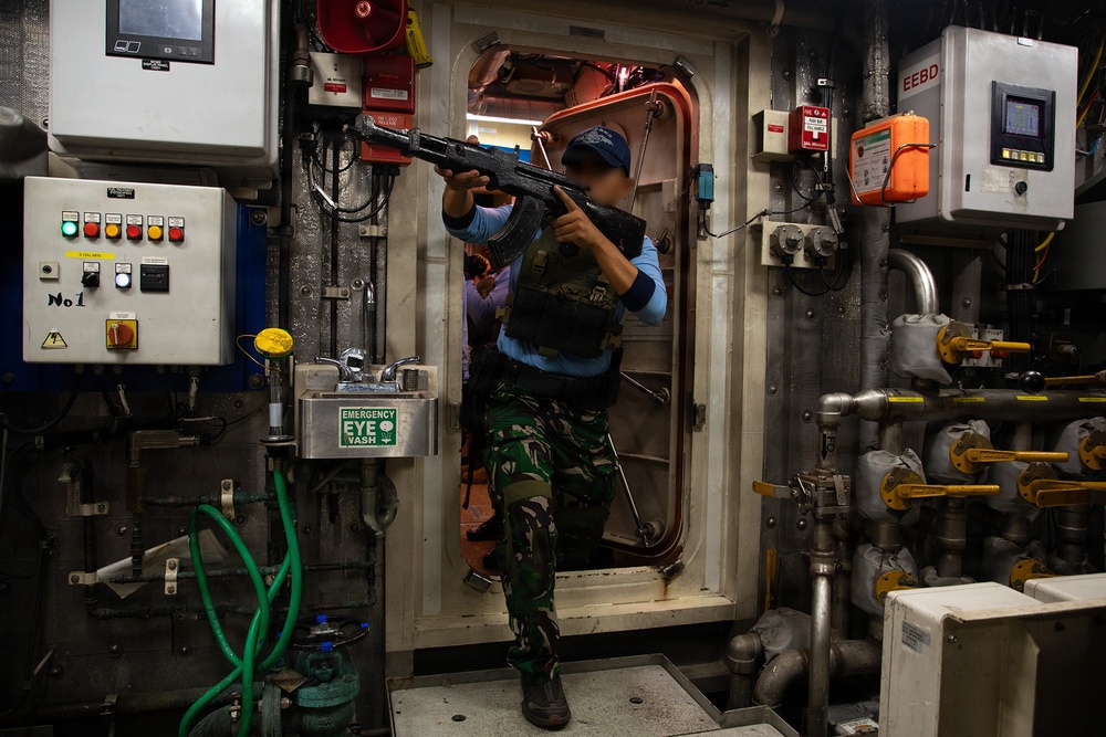 An armed individual wearing camouflage military fatigues, a tactical vest, and a blue cap is seen inside what appears to be a ship's engine room or mechanical compartment. The person is holding a rifle in a raised, ready position while standing in a doorway threshold. The setting contains industrial maritime equipment including control panels, piping, valves, and safety equipment.