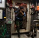 An armed individual wearing camouflage military fatigues, a tactical vest, and a blue cap is seen inside what appears to be a ship's engine room or mechanical compartment. The person is holding a rifle in a raised, ready position while standing in a doorway threshold. The setting contains industrial maritime equipment including control panels, piping, valves, and safety equipment.